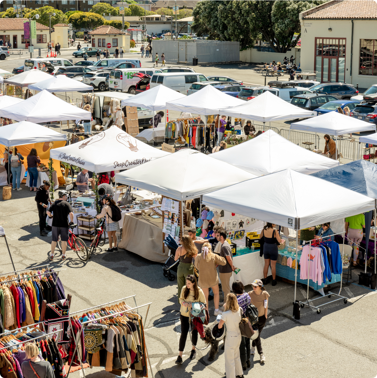 A sea of white tents and clothing racks fill the image, suggesting an outdoor farmer’s market or flea market where customers and sellers are meeting.