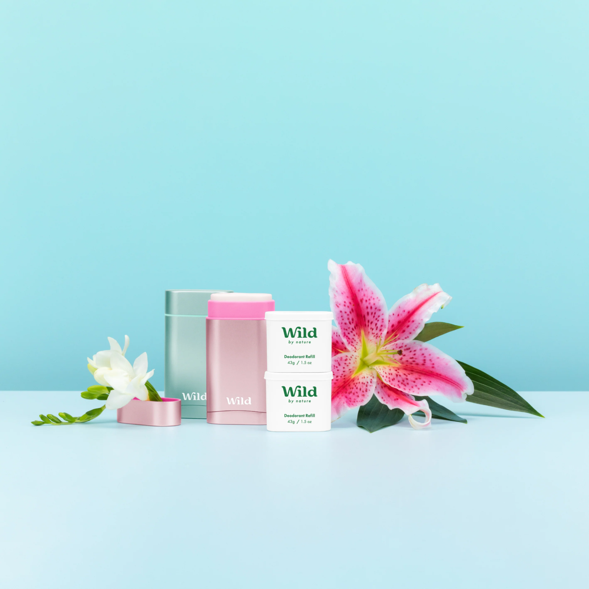 Pink and green deodorant sticks beside refills and tropical pink and white flowers. Wild Deodorant
