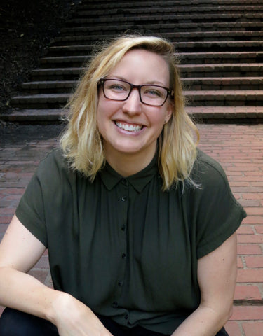 Beefcake Swimwear Founder Mel is sitting on outdoor stairs, smiling, wearing a green shirt and glasses.
